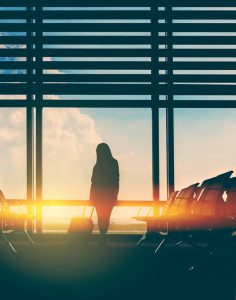 silgouette of woman in airport waiting for her flight, sunlight in background, carrying luggage.